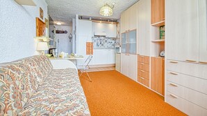 A compact living space with a patterned sofa, a dining table, a kitchen area with white cabinets, and a wooden wardrobe.