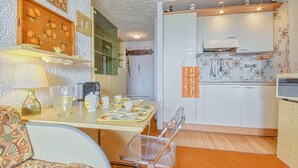 A compact kitchen with white cabinets, a built-in oven, and a microwave. There is a dining table with chairs, a lamp, and a coffee maker.