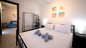 Ein Hotelzimmer mit einem ordentlich bezogenem Bett, einem kleinen Nachttisch mit Lampe, einem Wandregal und Blick auf den Flur.