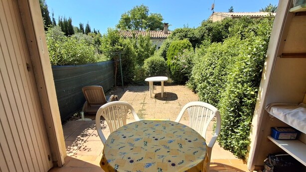 Un patio con una mesa redonda y dos sillas, rodeado de follaje y un suelo de baldosas.