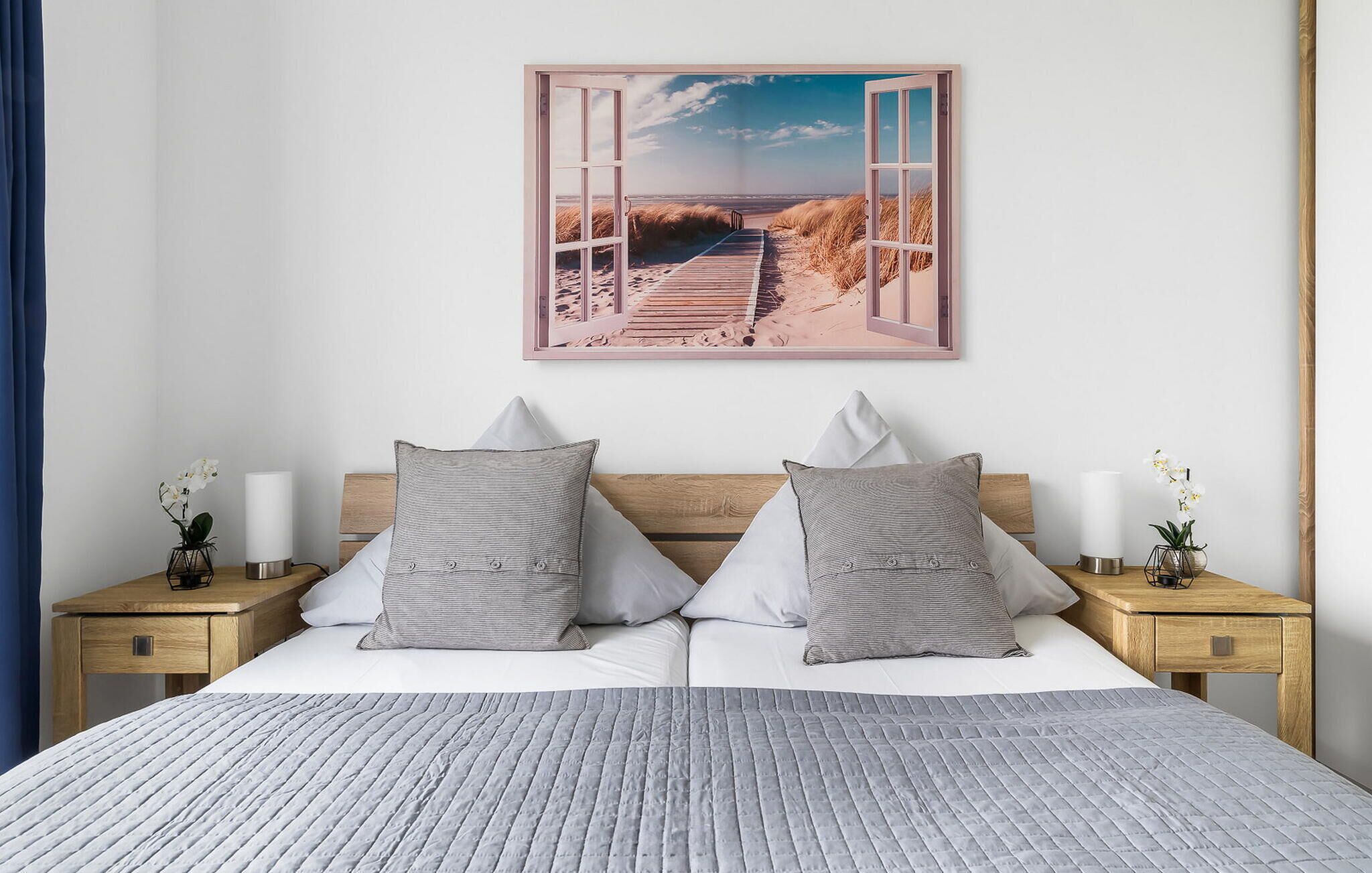 A bedroom with a bed, two nightstands, a wooden headboard, and a wall-mounted picture of a beach scene.