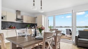 Une cuisine moderne avec vue sur l’eau, un coin repas avec une table dressée pour deux personnes et un canapé près de la fenêtre.