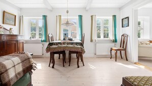 Een eetkamer met een houten tafel, stoelen en een kroonluchter. Er zijn ramen met gordijnen en uitzicht op groen buiten.