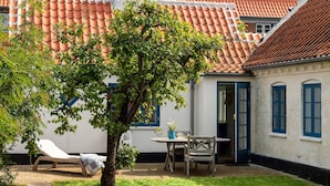 Een binnentuin met een boom, een ligstoel, een tafel met stoelen en een gebouw met rode dakpannen en blauwe kozijnen.