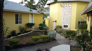 Ein gelbes Haus mit einer Steinmauer, ein kleiner Garten mit verschiedenen Pflanzen und einem Betonweg.
