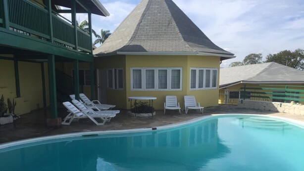 Ein Poolbereich mit einem gelben Gebäude, einer überdachten Terrasse und weißen Liegestühlen.