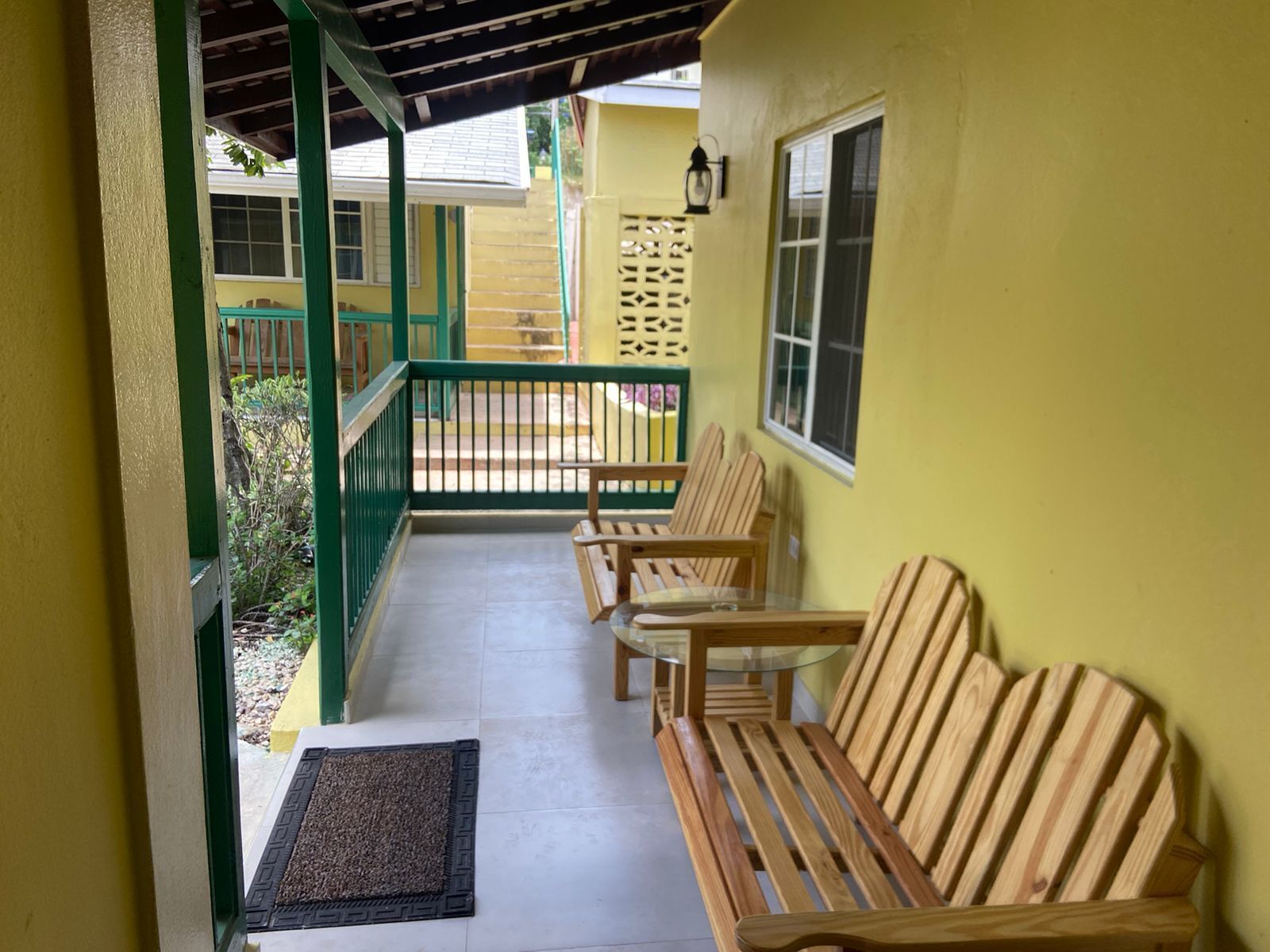 Une véranda avec des bancs en bois et une table en verre, un mur jaune et une rambarde verte.