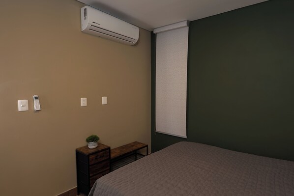 A bedroom with a bed, an air conditioner, a bedside table with a plant, a bench, and a window with blinds.
