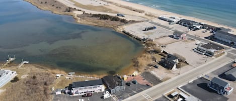 An aerial view of a coastal area with a large body of water, a beach, and various buildings including a hotel with a parking lot and a road.
