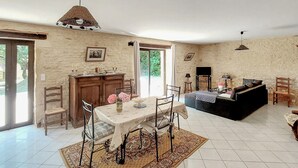 A spacious living area with a dining table, chairs, a sofa, and a TV. There is a large window providing natural light and a view of greenery outside. - Lovely home in Paulin with kitchen (Paulin)
