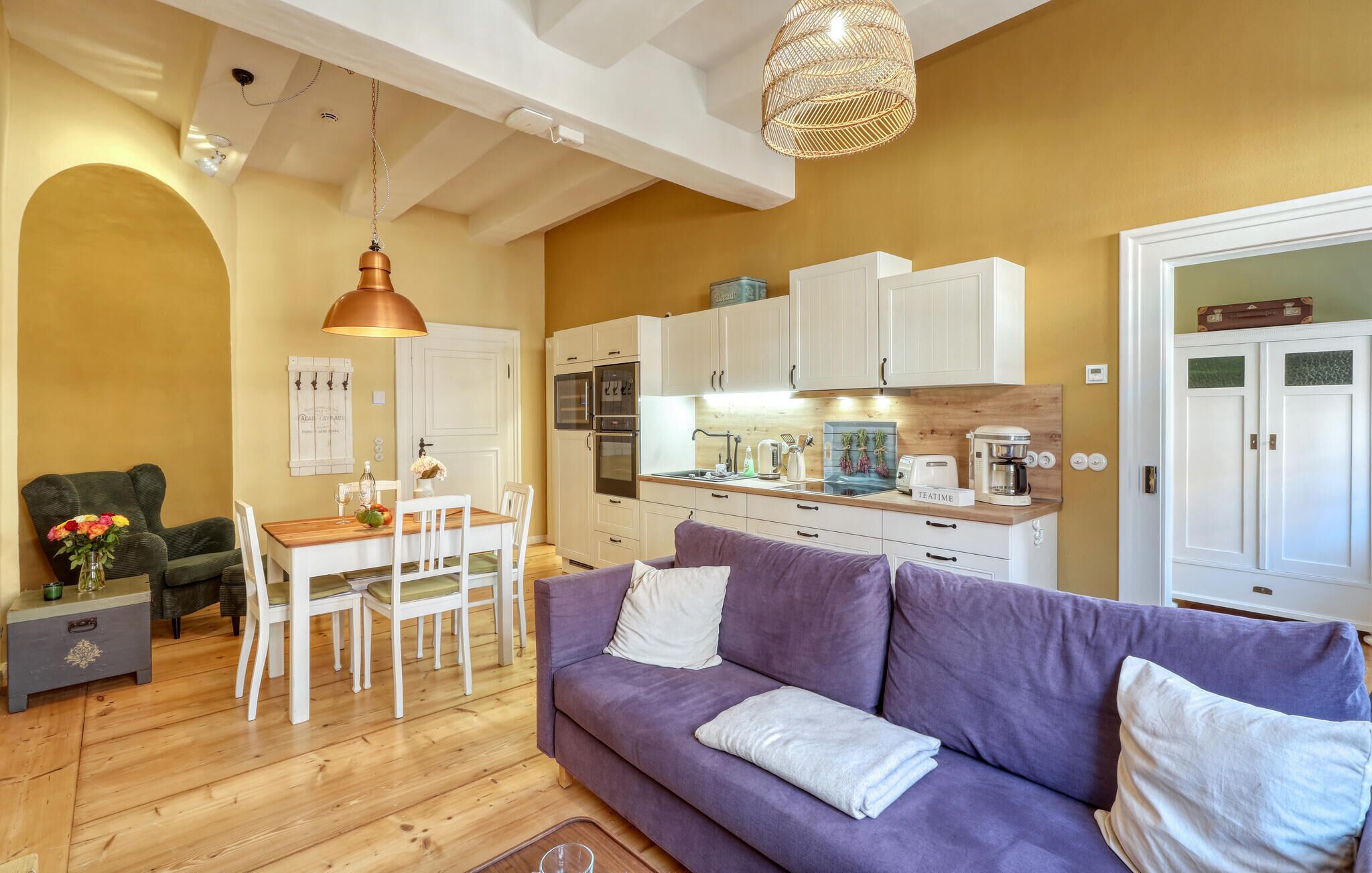 Un salon chaleureux avec un canapé violet, une table à manger en bois et des chaises. On peut apercevoir une cuisine avec des meubles blancs et une machine à café.