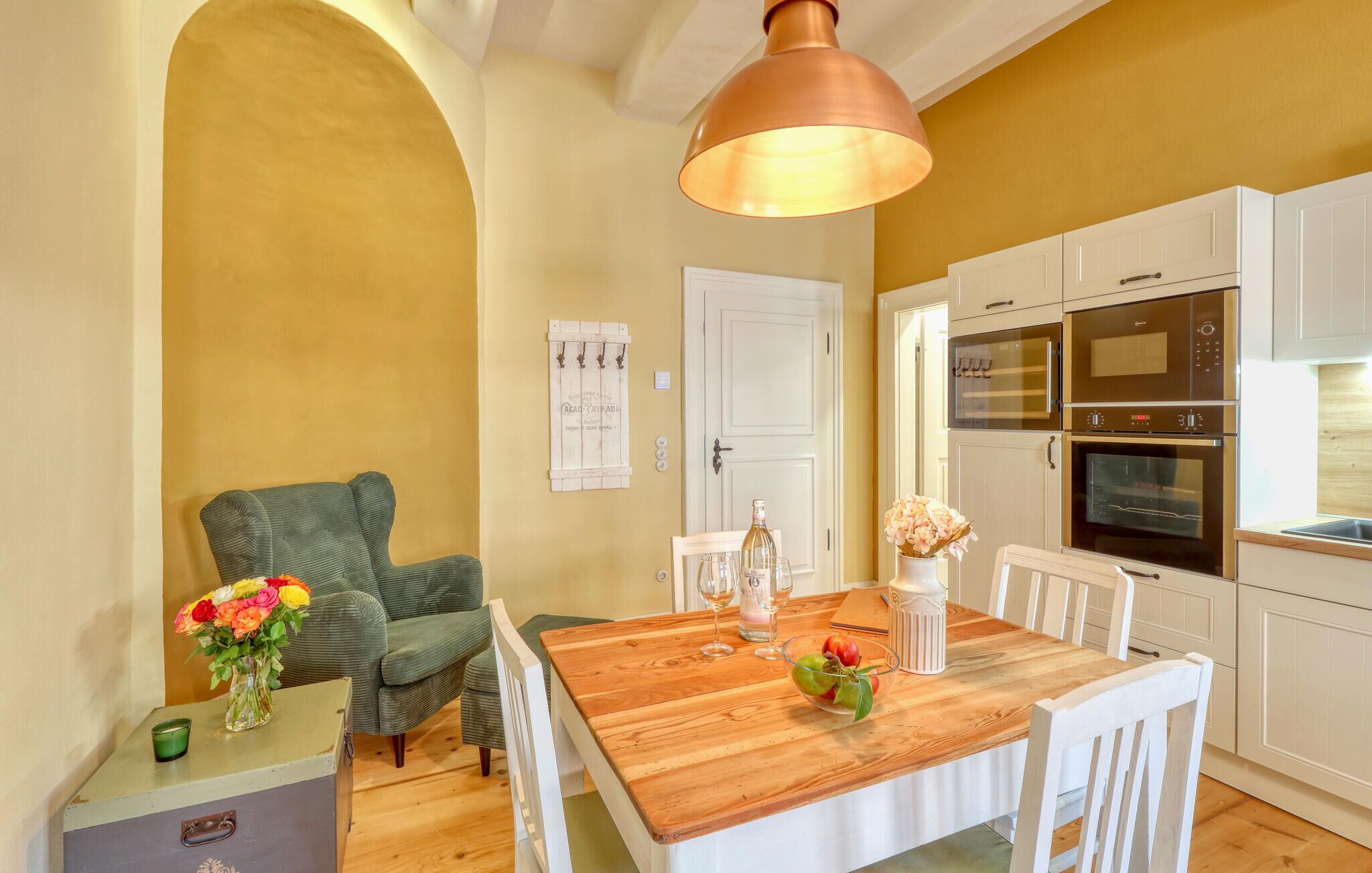 Un coin repas avec une table en bois, des chaises blanches, un fauteuil vert et une cuisine dotée de meubles blancs et d’appareils électroménagers en acier inoxydable.