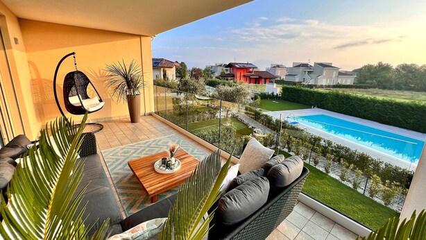 Un balcón con una silla colgante, una mesa de madera y un sofá. Se ve una piscina al fondo.