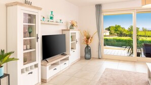 Una sala de estar moderna con una televisión de pantalla plana, un mueble con puertas de vidrio, un jarrón grande con flores secas y vistas a un paisaje verde a través de una puerta corrediza de vidrio.
