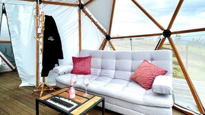 A tent with a white interior, a grey sofa with red cushions, a wooden coffee table, and a black jacket hanging on a rack. - Glamping Spa con vista embalse de Tominé (Guatavita)