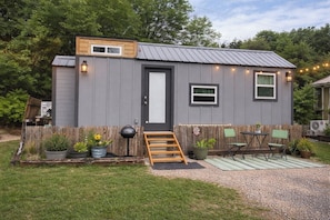 A small, single-story building with a front door, a window, and a small porch area with a chair and table. - Shangri-La Tiny Home with WiFi near Winery, Lakes, and Antique Shops!! (Azle)