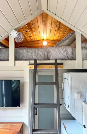 A loft-style bedroom with a wooden ceiling, a ladder leading to a bunk bed, a washing machine, and a flat-screen TV. - Shangri-La Tiny Home with WiFi near Winery, Lakes, and Antique Shops!! (Azle)