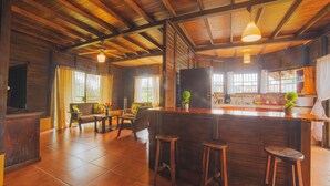 Un'ampia cucina con una zona bar in legno, un tavolo da pranzo con sedie e un divano in un soggiorno con ampie finestre.