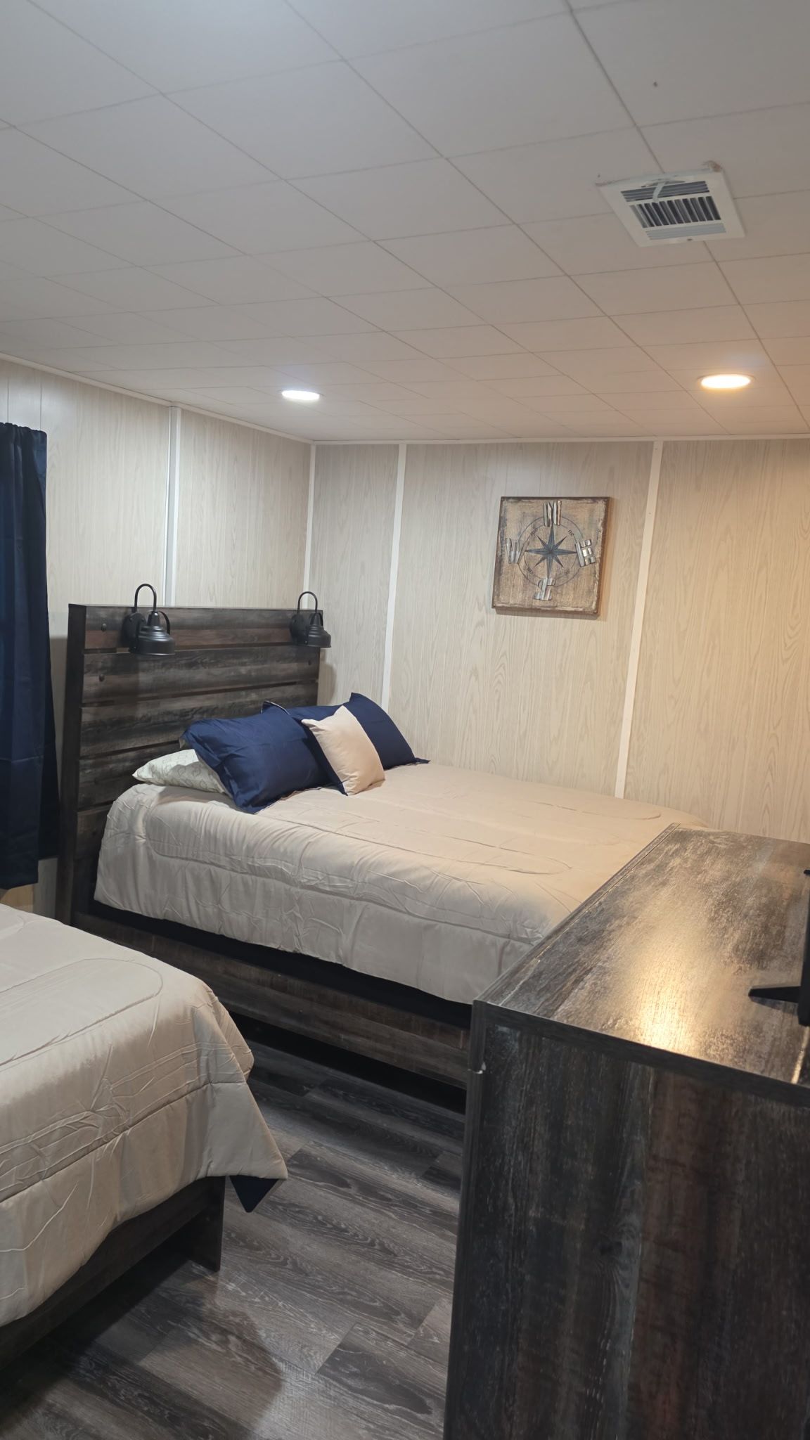A room with a bed, a bedside table, a headboard, a wall clock, and a ceiling vent.