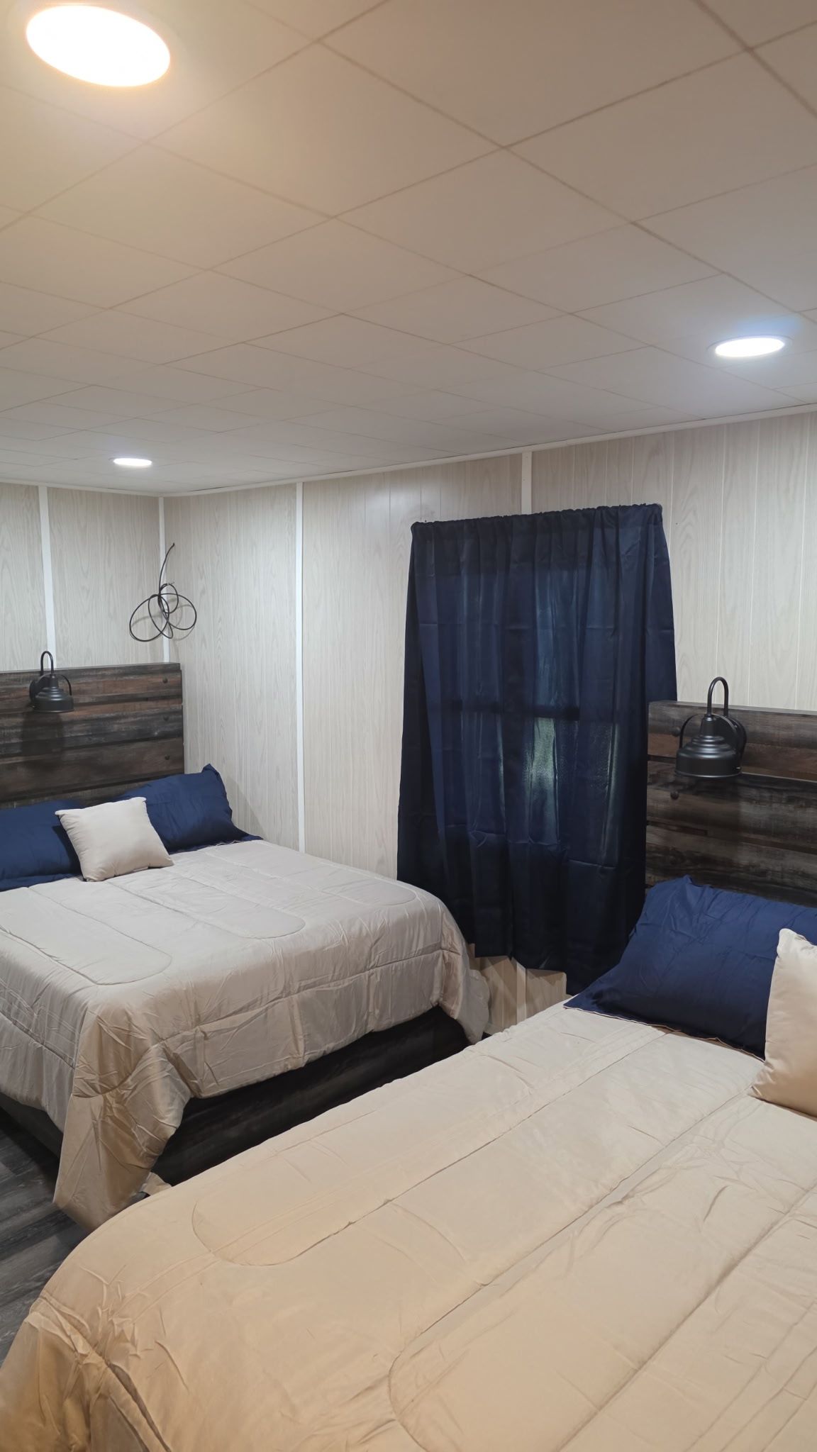 Habitación de hotel con dos camas, cabecero de madera, cortina oscura y una tetera en la mesita de noche.