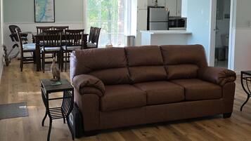 A living room with a brown sofa, wooden flooring, a dining area with chairs, a kitchen with appliances, and a ceiling fan.