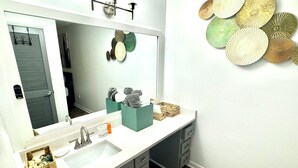 Ein Badezimmer mit einem großen Spiegel, einem Waschbecken und einem Waschtisch mit Schubladen. Auf dem Waschtisch stehen dekorative Gegenstände und über dem Spiegel ist eine Wanddekoration angebracht.