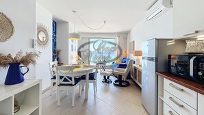 A modern kitchen with white cabinets, a microwave, and a Samsung refrigerator. There is a dining area with a white table and chairs, and a blue vase with dried plants on the left.