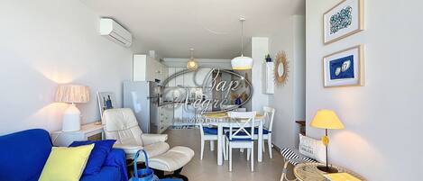 A modern living room with a blue sofa, a dining area with white chairs, and a kitchen in the background.
