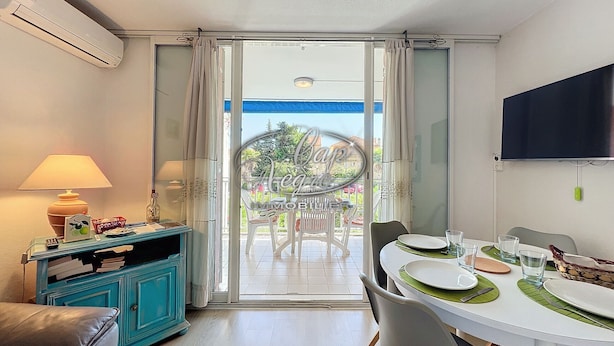 A dining area with a table set for two, a television mounted on the wall, and a view of the outdoors through a glass door.