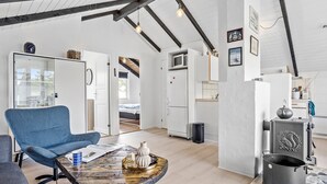 Een ruime woonkamer met een hoge plafond, zichtbare balken, een groot raam en een keuken met een fornuis en een koelkast.