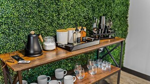 Ein Holztisch mit Kaffeemaschine, Milchkrug und Kaffeetassen, vor einer grünen, blätterreichen Wand.