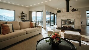 A modern living room with a sofa, a coffee table with flowers, a fireplace, and large windows offering a view of mountains. - The Heights  (Wanaka)