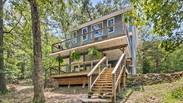 Une cabane en bois à deux étages, dotée d’un escalier en pierre et d’un balcon, et entourée d’arbres.