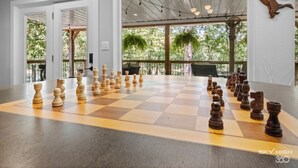 Un grand jeu d’échecs en bois est installé dans une pièce spacieuse, dotée de grandes fenêtres et offrant une vue sur les arbres à l’extérieur.