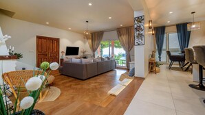 Een moderne woonkamer met een grijze bank, houten vloeren, een flatscreen televisie en een eethoek met een tafel en stoelen.
