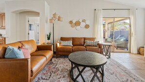 Ein geräumiges Wohnzimmer mit einer braunen Ledersofa, einem runden Couchtisch und einem gemusterten Teppich. Es gibt einen Essbereich mit einem kleinen Tisch und Stühlen sowie eine große Schiebeglasdoor, die zu einer Außenpatio führt.