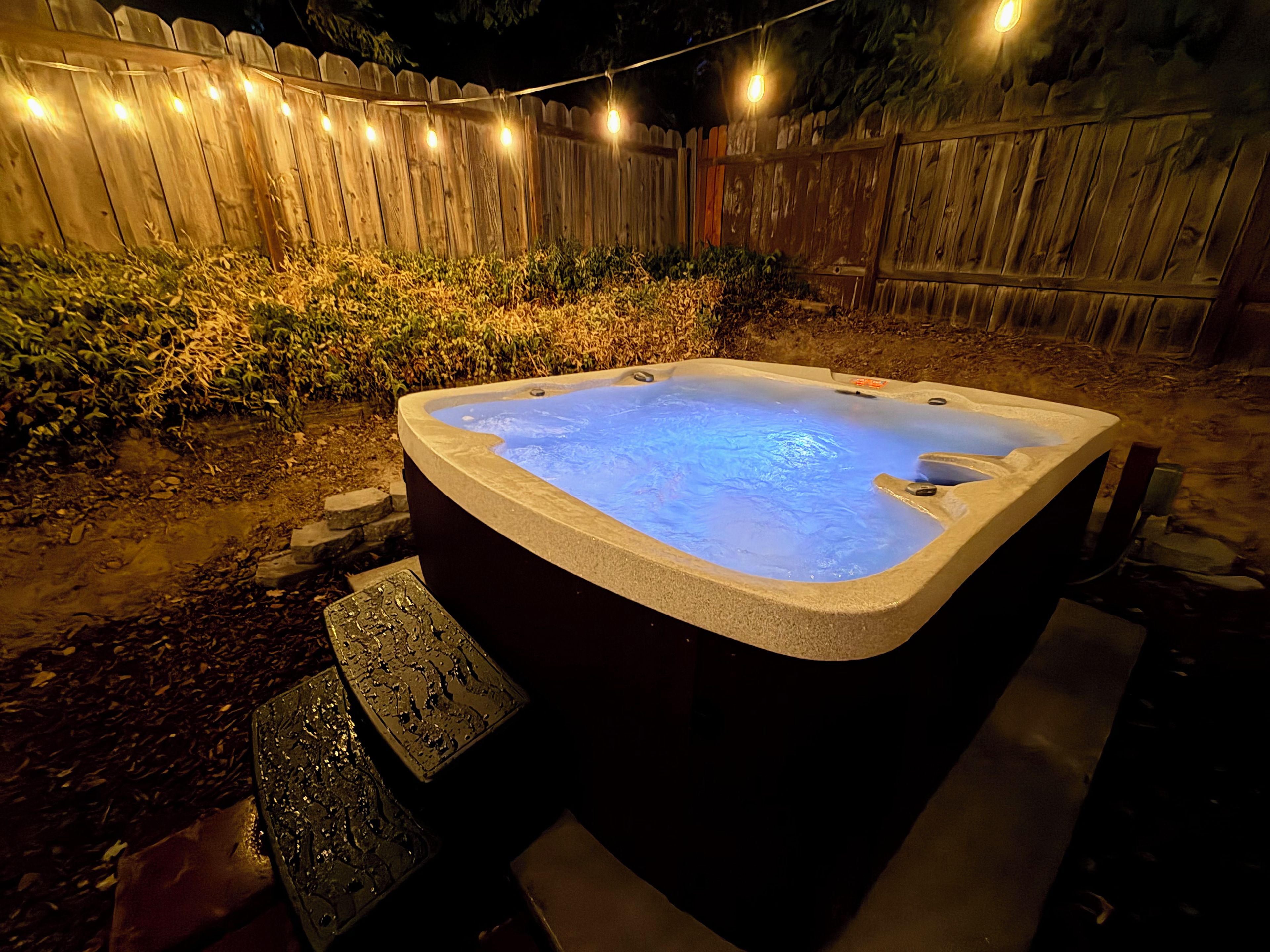 A hot tub in a backyard with a wooden fence and string lights.