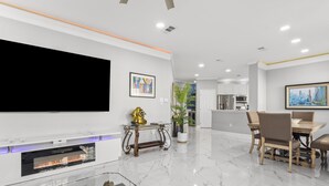 A modern living room with a flat-screen TV, a fireplace, a dining area with a table and chairs, and a kitchen in the background.
