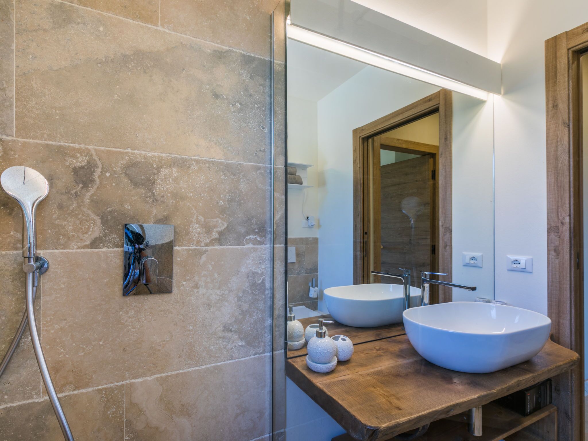 A modern bathroom with two large white sinks, a glass shower enclosure, and a wooden vanity.