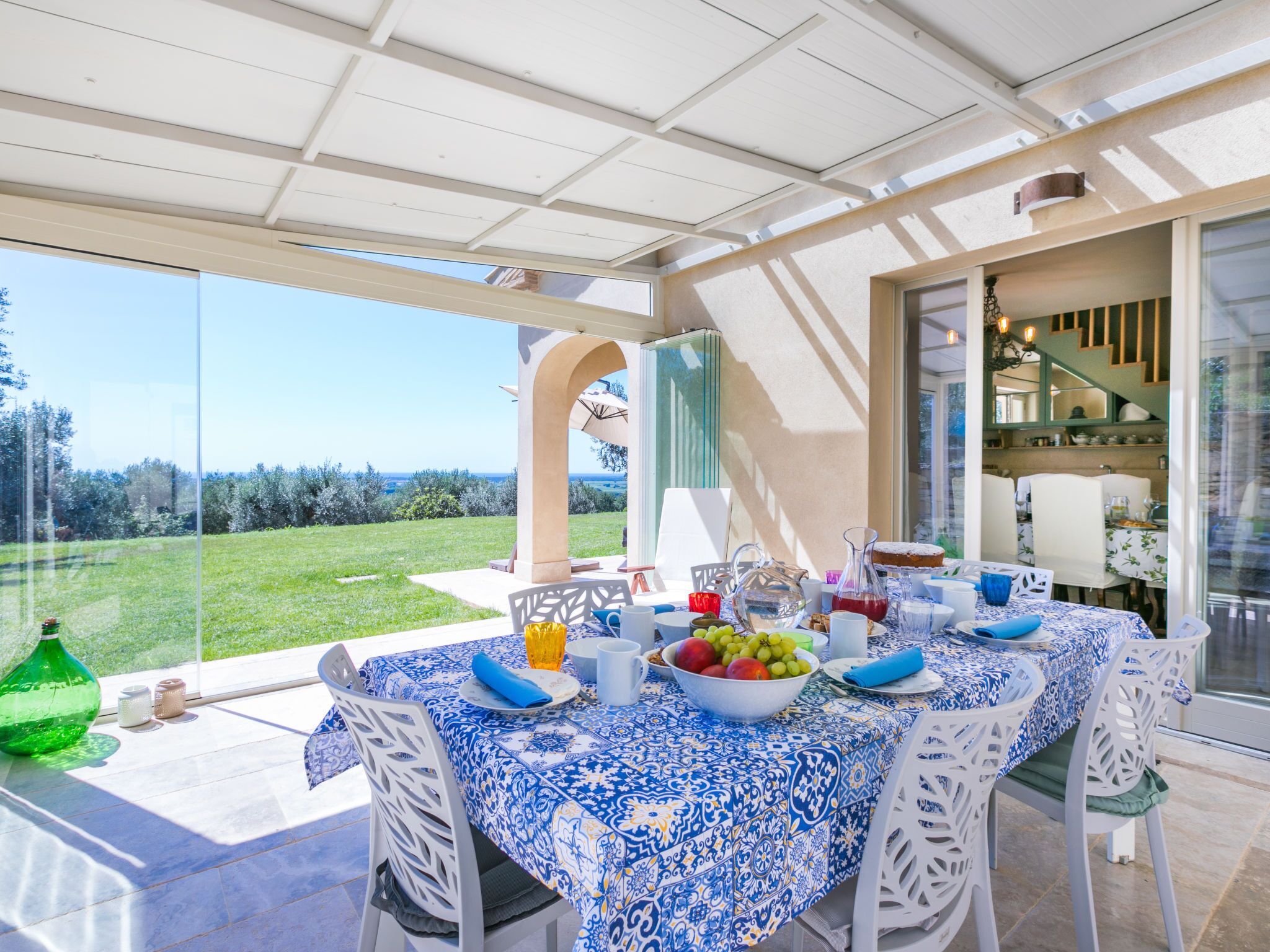 An outdoor dining area with a table set for four, surrounded by white chairs, a patterned tablecloth, and a view of the countryside.
