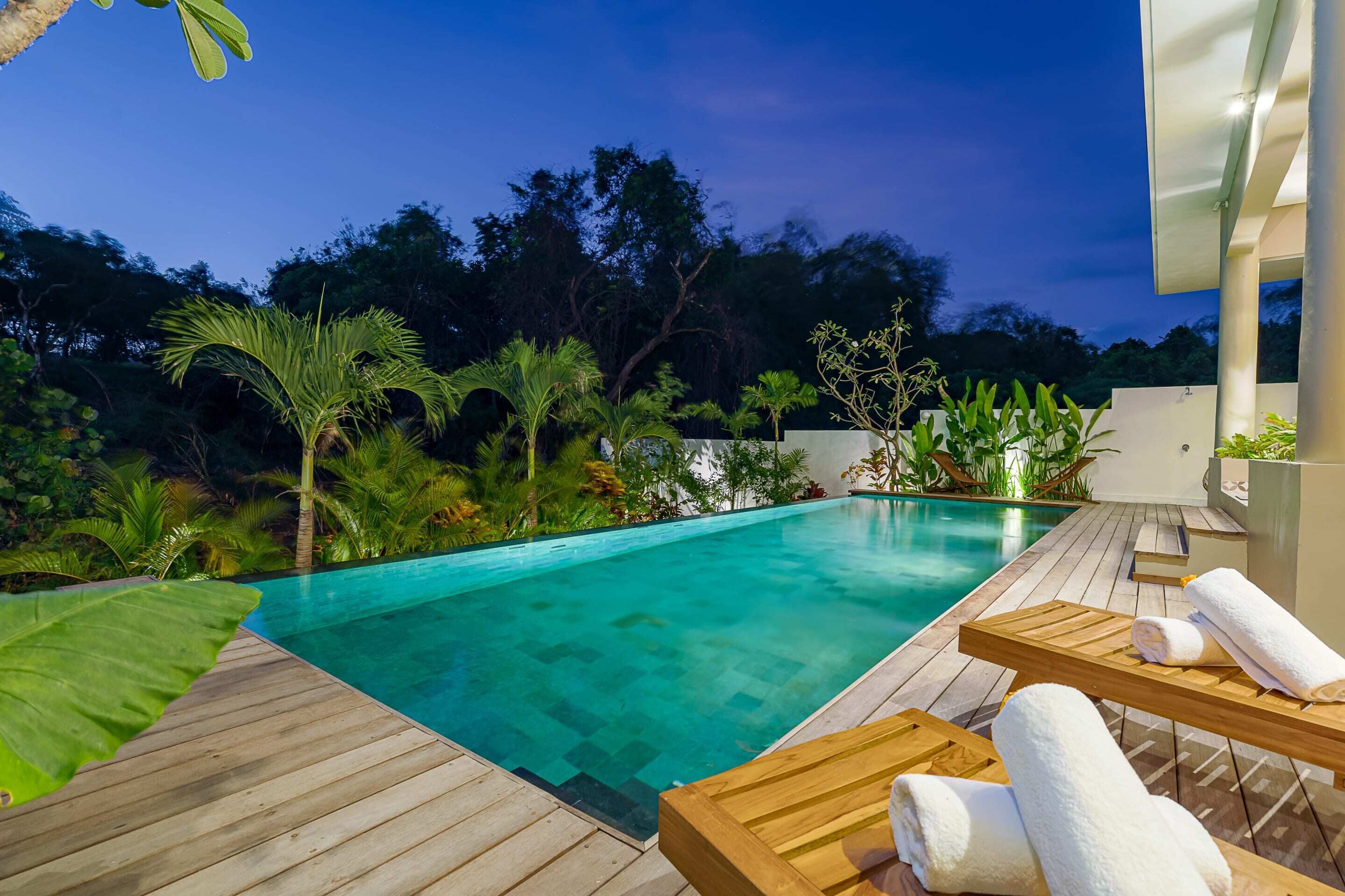 Una terraza de madera con piscina, rodeada de plantas tropicales y un edificio moderno.