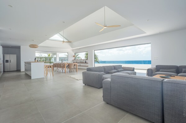 A spacious living area with a large window overlooking the ocean, a modern kitchen, and a dining area. - La Plage Martinique (Schoelcher)