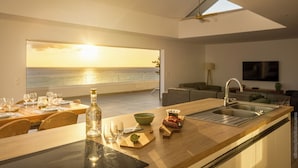 Une cuisine moderne avec vue sur l’océan, un coin repas avec une table dressée, et un espace de vie comprenant un canapé et un téléviseur.