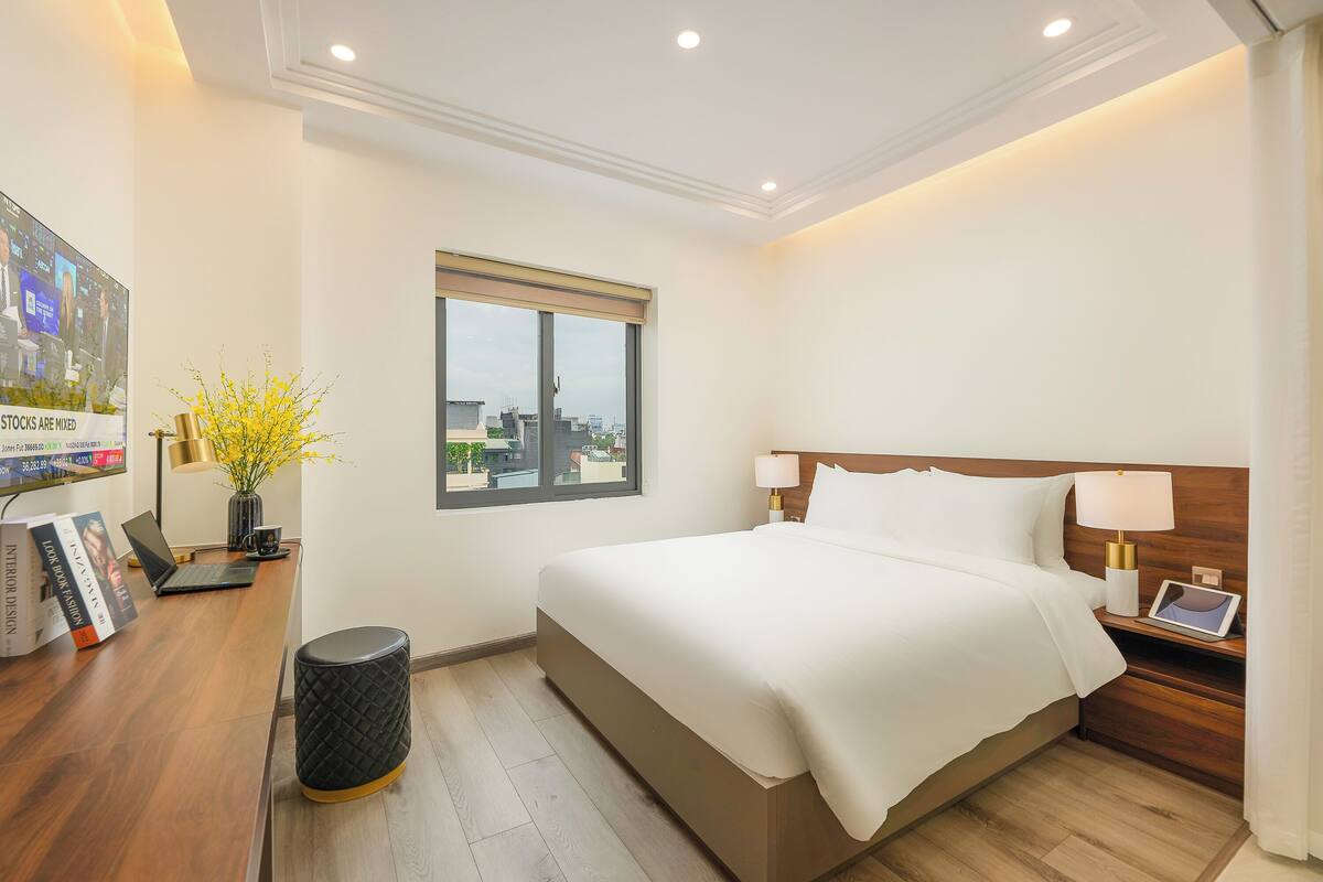 A hotel room with a large bed, a desk with a laptop, a vase with yellow flowers, and a window with a city view.