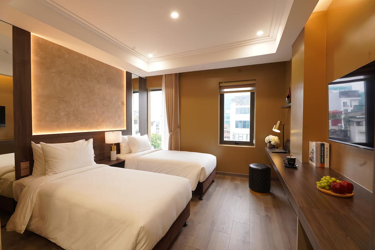 A hotel room with two beds, a wooden floor, a desk with a fruit bowl, and a window with a view of buildings.