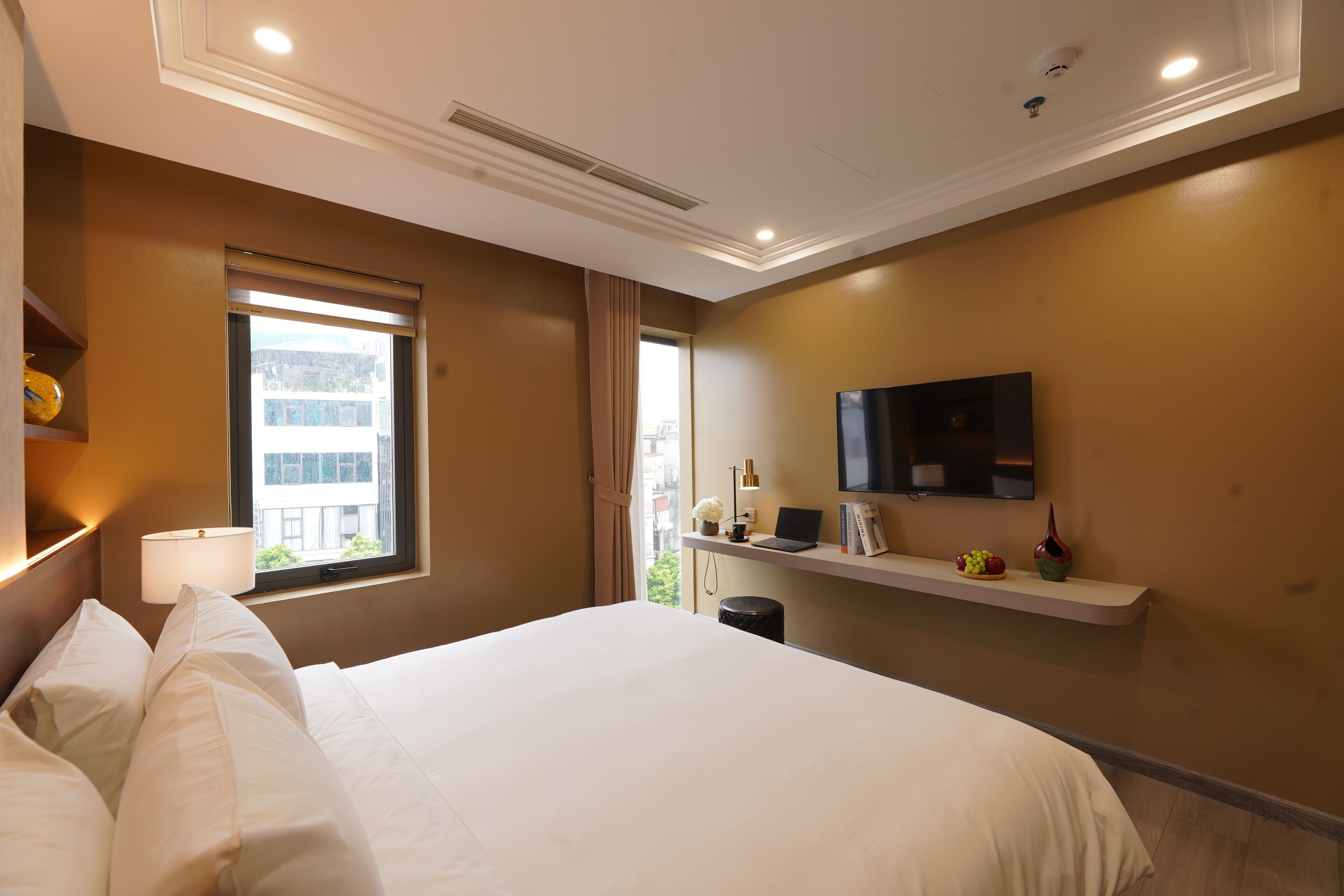 A hotel room with a large bed, a TV mounted on the wall, a window with a view of buildings, and a small desk with a lamp and a laptop.