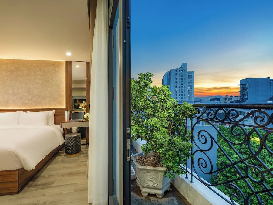 Quarto de hotel com uma cama grande, mesa de cabeceira, luminária e planta em vaso.