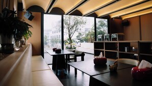 A modern interior with a wooden table, chairs, a bookshelf, and a large window offering a view of the outdoors. -  Gilson Hanoi Hotel (Hanoi)