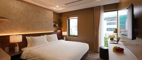A hotel room with a large bed, bedside lamps, a small table with fruit, a window with curtains, and a city view.