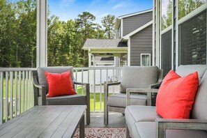A covered outdoor seating area with a table and two chairs, a railing, and a view of a house in the background.